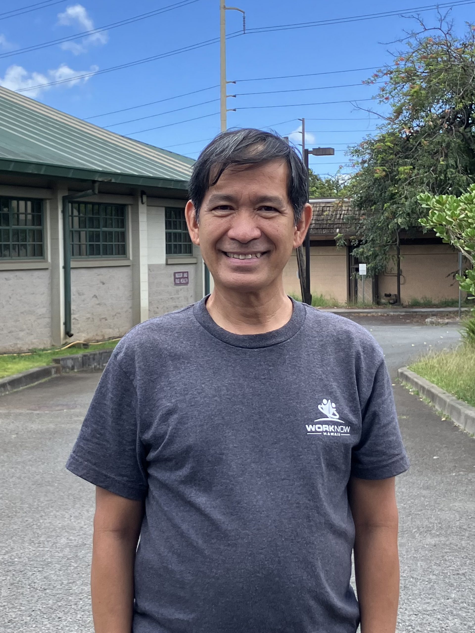 Image of Hernani standing outside a building, in his Work Now Hawaii shirt."